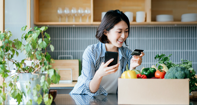 Image to support the blog. A lady in her kitchen smiling while doing online shopping on her phone and/or paying with her credit card.